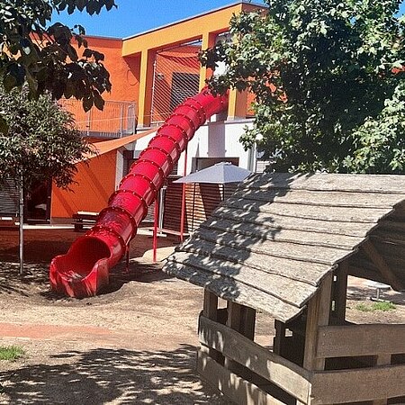 Farbenfroher Außenbereich der Kita Schaukel mit einem großen roten Röhrenrutschenturm, umgeben von Bäumen und Sträuchern. Im Vordergrund steht ein kleines Holzhäuschen, und der Boden ist teils mit Sand, teils mit Wegen gestaltet. Die Umgebung wirkt einladend und kinderfreundlicharbenfroher Außenbereich eines Kindergartens mit einem großen roten Röhrenrutschenturm, umgeben von Bäumen und Sträuchern. Im Vordergrund steht ein kleines Holzhäuschen, und der Boden ist teils mit Sand, teils mit Wegen gestaltet. Die Umgebung wirkt einladend und kinderfreundlich