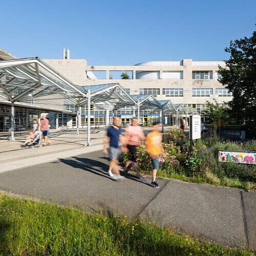 Außenansicht eines modernen Gebäudes mit Glasüberdachungen und Gehwegen. Menschen spazieren bei sonnigem Wetter entlang des Klinik-Eingangsbereichs, umgeben von grünen Pflanzen und Blumen