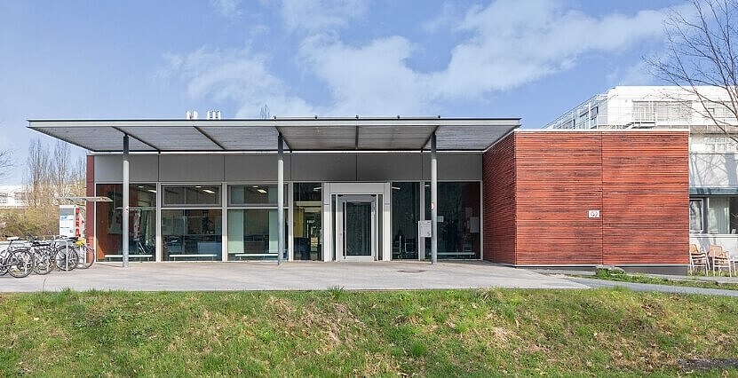 Flaches Gebäude des A.R.Z. mit moderner Architektur, Holz- und Glaselementen. Vor dem Gebäude stehen Fahrräder und ein Schild mit der Aufschrift „ARZ“. Im Vordergrund ist eine kleine Grünfläche zu sehen