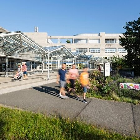 Außenansicht eines modernen Gebäudes mit Glasüberdachungen und Gehwegen. Menschen spazieren bei sonnigem Wetter entlang des Klinik-Eingangsbereichs, umgeben von grünen Pflanzen und Blumen