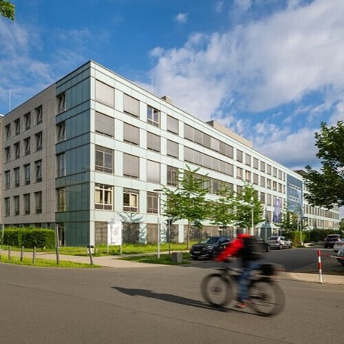 Ein mehrstöckiges Klinikgebäude mit einer klaren, modernen Architektur und großen Glasflächen. Vor dem Gebäude befinden sich geparkte Autos und ein Radfahrer fährt auf der Straße vorbei. Der Himmel ist blau mit einigen Wolken