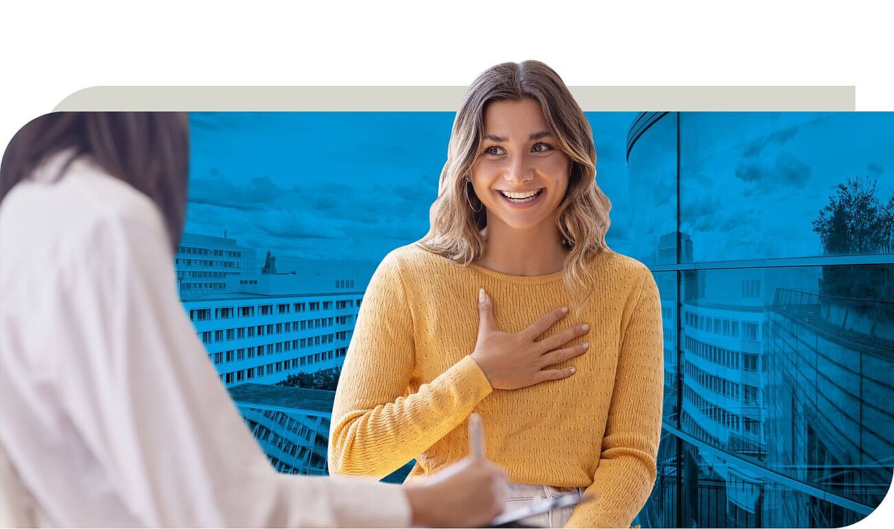 Eine Frau in einem gelben Pullover sitzt und hält eine Hand auf ihrer Brust, während sie mit einer anderen Person spricht, die im Vordergrund von hinten zu sehen ist. Der Hintergrund zeigt eine moderne Glasfassade mit blauem Farbton und Gebäudestrukturen.