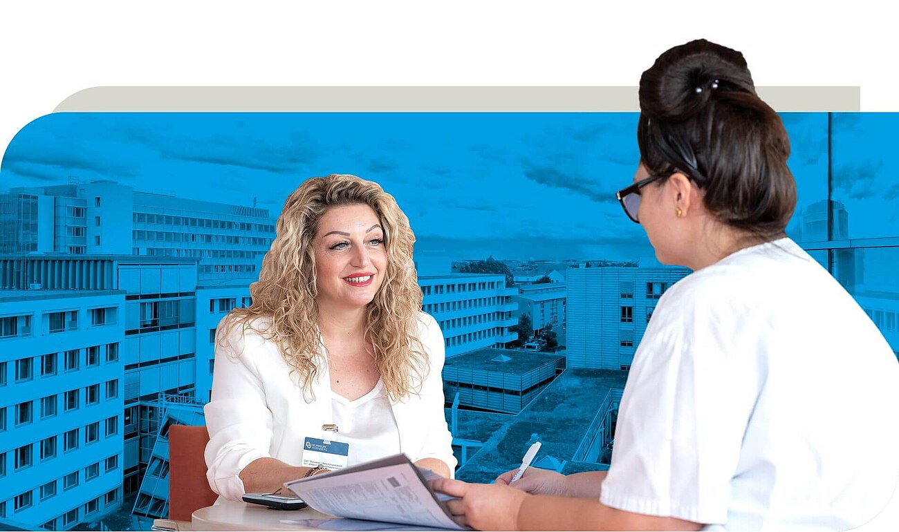 Zwei Frauen sitzen an einem Tisch in einem Gespräch. Eine Frau trägt ein weißes Oberteil und hält Dokumente, während die andere Frau in Arbeitskleidung Notizen macht. Das Bild stellt ein Beratungsgespräch dar