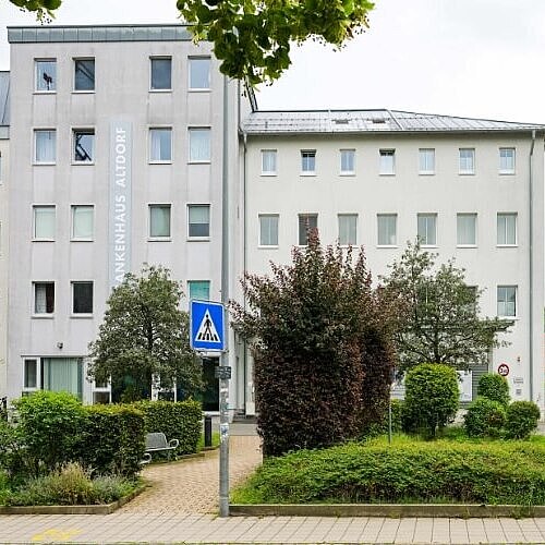 Außenansicht des Altdorfer Krankenhausgebäudes mit der Aufschrift „Krankenhaus“ an der Fassade. Ein Glasvordach markiert den Eingangsbereich, umgeben von Bäumen und Sträuchern