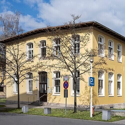 Gebäude mit klassischer Architektur auf dem Gelände des Campus Nord des Klinikums Nürnberg, umgeben von Bäumen und einer gepflegten Grünfläche. Im Vordergrund befinden sich eine Uhr und ein Parkplatzschild