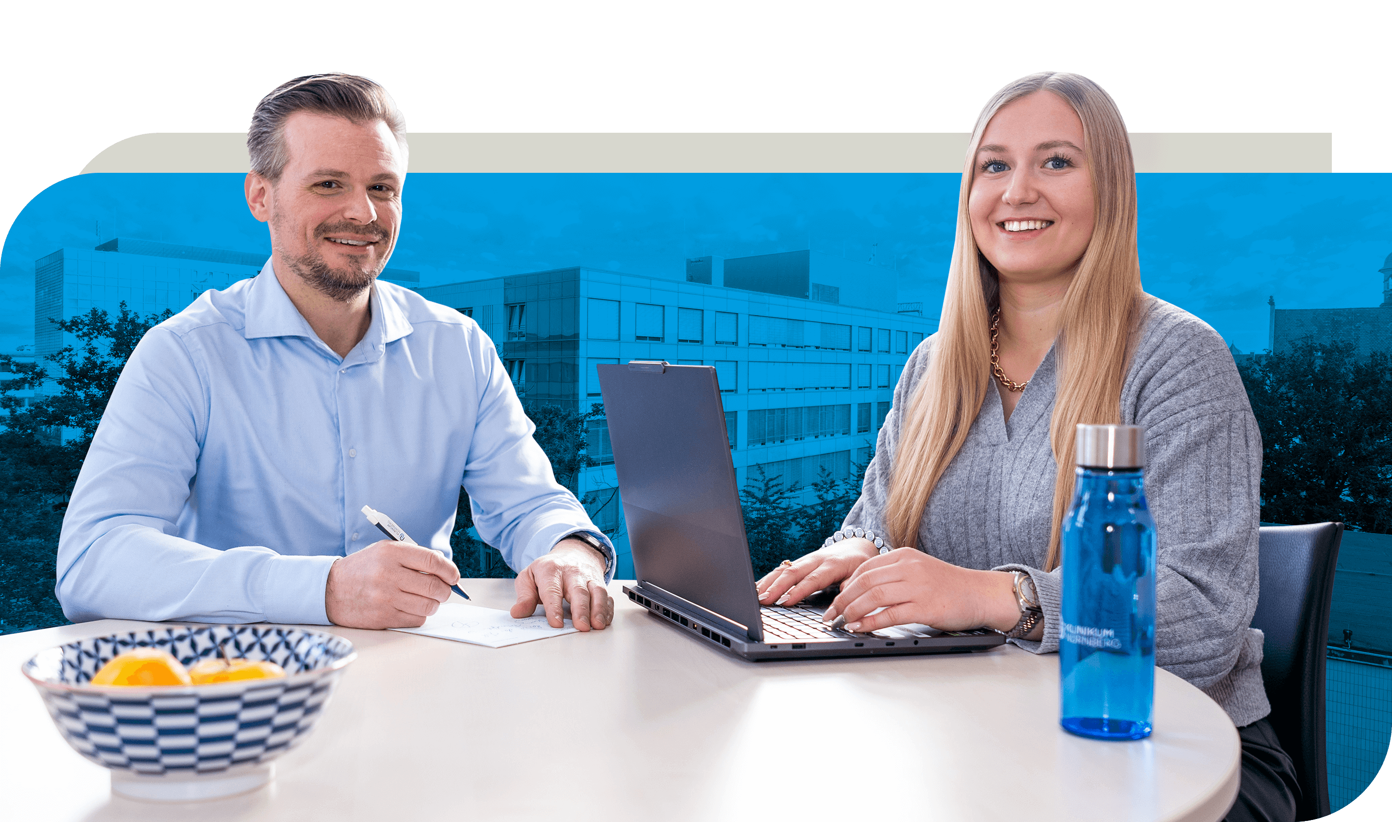 Zwei Personen sitzen an einem Tisch. Eine Person schreibt Notizen auf Papier, während die andere an einem Laptop arbeitet. Auf dem Tisch stehen eine blaue Wasserflasche und eine Schale mit Obst. Im Hintergrund ist ein Bürogebäude zu erkennen
