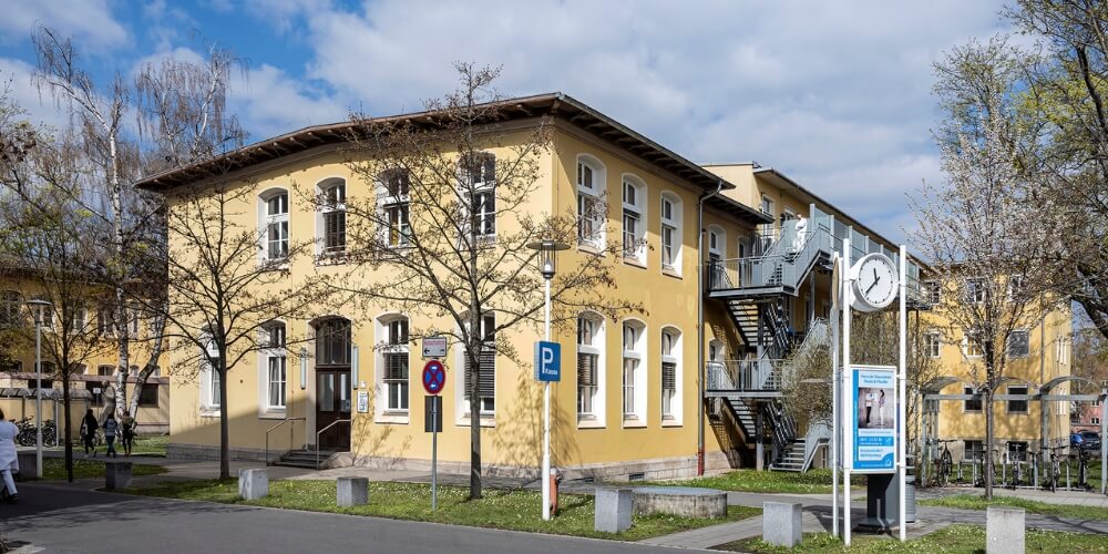 Gebäude mit klassischer Architektur auf dem Gelände des Campus Nord des Klinikums Nürnberg, umgeben von Bäumen und einer gepflegten Grünfläche. Im Vordergrund befinden sich eine Uhr und ein Parkplatzschild