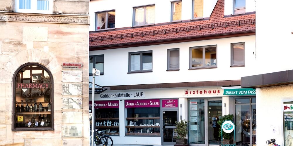 Ärztehaus in einem historischen Gebäude mit einem Schaufenster, das „Pharmacy“ zeigt. Daneben ein moderner Gebäudeteil mit der Aufschrift „Ärztehaus“ sowie Geschäften