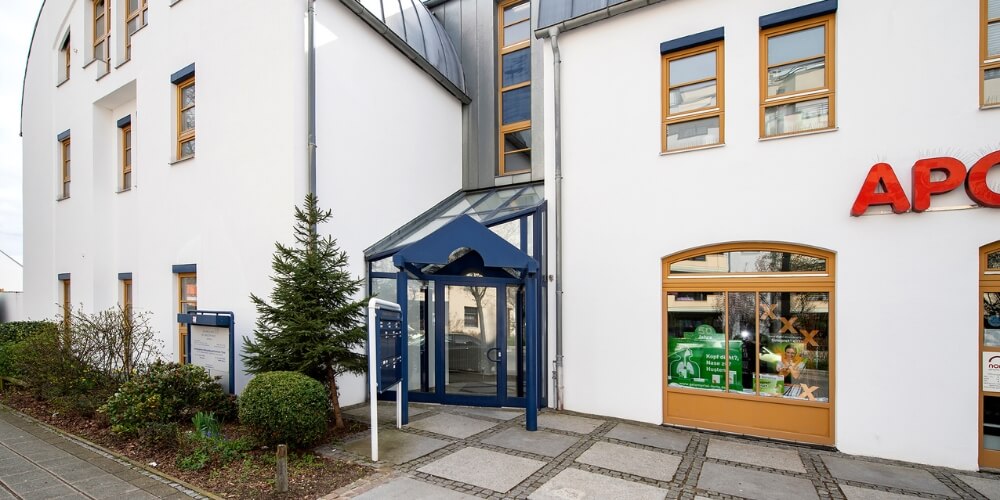 Eingangsbereich des ABC Thon in weißen Gebäudes mit blauen Fensterrahmen und einem gläsernen Vordach. Rechts im Bild befindet sich eine Apotheke mit rotem Schriftzug