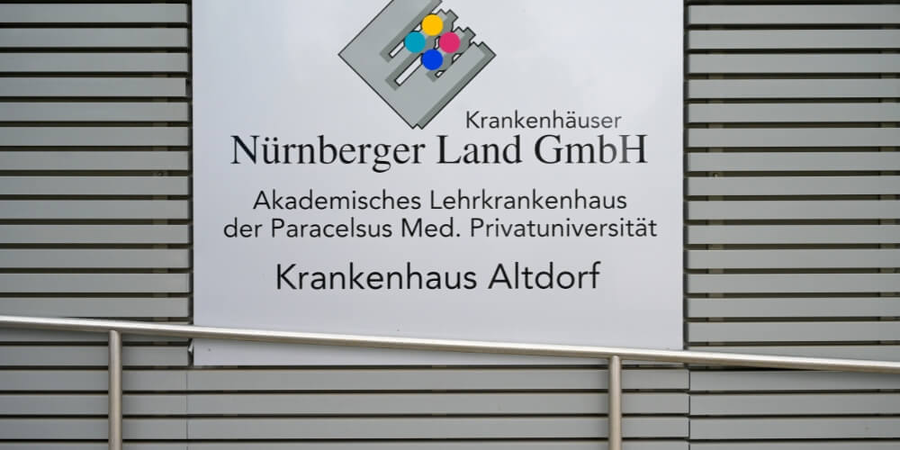 Nahaufnahme eines Schildes am Krankenhaus Altdorf mit der Aufschrift: ‚Krankenhäuser Nürnberger Land GmbH – Akademisches Lehrkrankenhaus der Paracelsus Med. Privatuniversität – Krankenhaus Altdorf