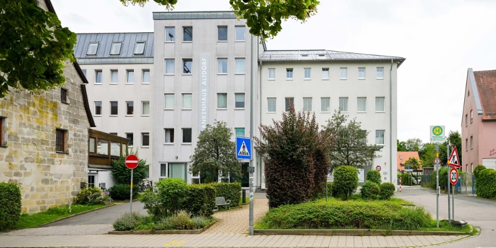 Außenansicht des Altdorfer Krankenhausgebäudes mit der Aufschrift „Krankenhaus“ an der Fassade. Ein Glasvordach markiert den Eingangsbereich, umgeben von Bäumen und Sträuchern