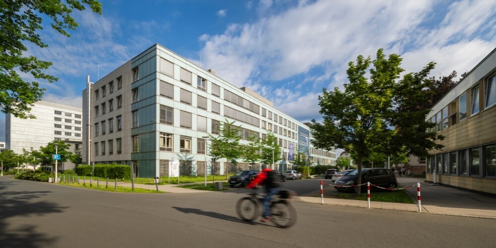 Ein mehrstöckiges Klinikgebäude mit einer klaren, modernen Architektur und großen Glasflächen. Vor dem Gebäude befinden sich geparkte Autos und ein Radfahrer fährt auf der Straße vorbei. Der Himmel ist blau mit einigen Wolken