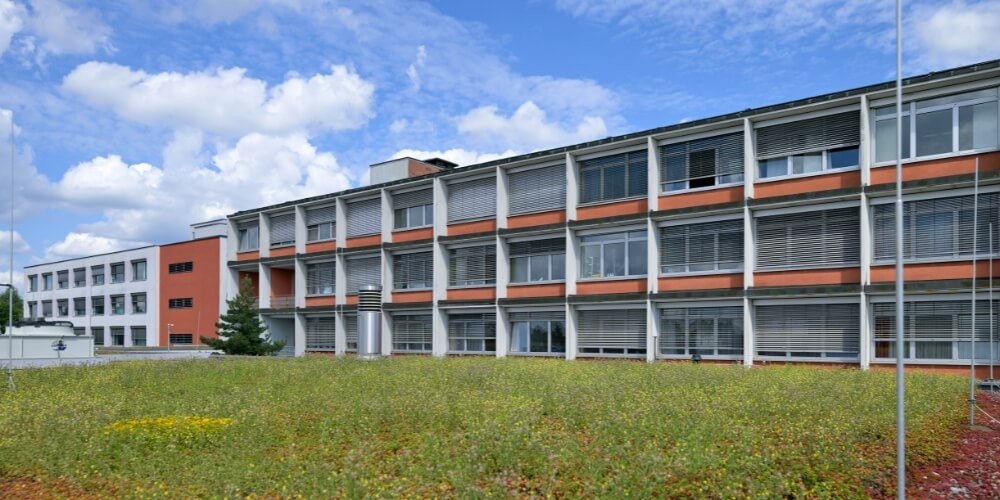 Außenansicht des Krankenhauses Lauf mit großen Fenstern und Sonnenschutzlamellen. Im Vordergrund ist ein begrüntes Dach mit Wildblumen zu sehen, darüber ein blauer Himmel mit weißen Wolken