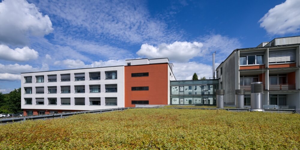 Modernes Gebäude des Krankenhauses Lauf mit weißer und roter Fassade, verbunden durch einen gläsernen Übergang. Im Vordergrund ist ein bepflanztes Gründach zu sehen, darüber ein blauer Himmel mit weißen Wolken