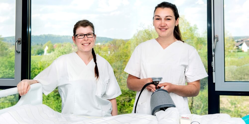 Zwei Pflegekräfte in weißer Arbeitskleidung stehen vor einem großen Fenster mit Blick auf eine grüne Landschaft. 