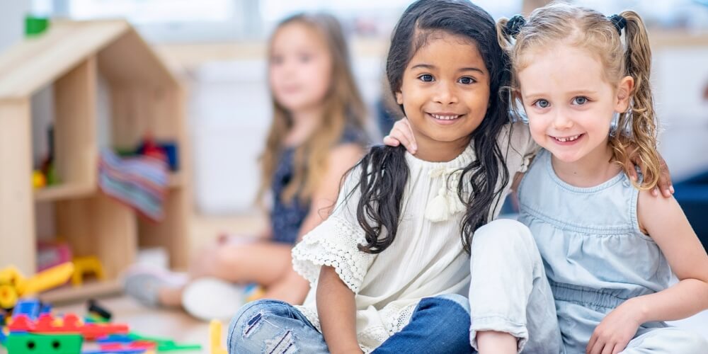 Zwei Kinder sitzen eng beieinander in einer hellen Kindertagesstätte. Im Hintergrund sind Holzspielzeug und weitere Kinder zu sehen