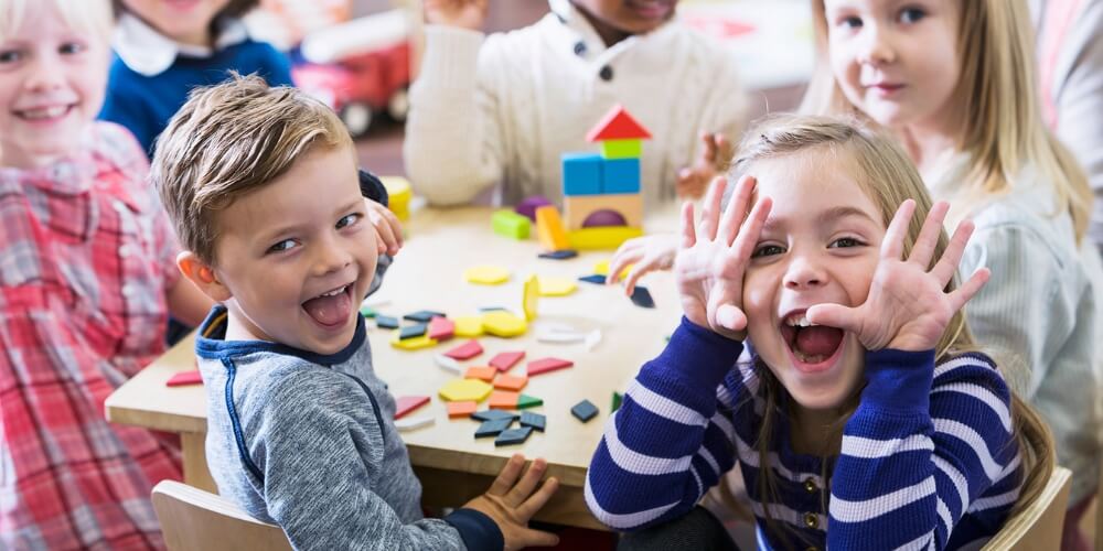 Kinder sitzen an einem Tisch und spielen mit bunten Bauklötzen und geometrischen Formen. Die Szene zeigt eine fröhliche Atmosphäre in einer Kindertagesstätte