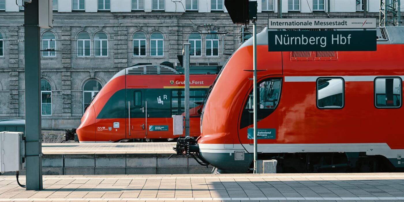 Zwei rote Regionalzüge stehen nebeneinander an einem Bahnsteig des Nürnberger Hauptbahnhofs. Im Vordergrund ist ein Schild mit der Aufschrift „Nürnberg Hbf“ zu sehen. Im Hintergrund befindet sich ein historisches Gebäude mit großen Fenstern. Auf einem der Züge ist der Schriftzug „Grüß Gott. Forst“ sichtbar, während der andere mit „Bahnland Bayern“ beschriftet ist.