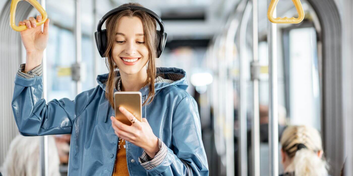 Junge Frau in einer blauen Jacke mit Kopfhörern, die in öffentlichen Verkehrsmitteln steht und lächelnd auf ihr Smartphone schaut. Symbolisiert Mobilitätsvorteile wie ein Jobticket