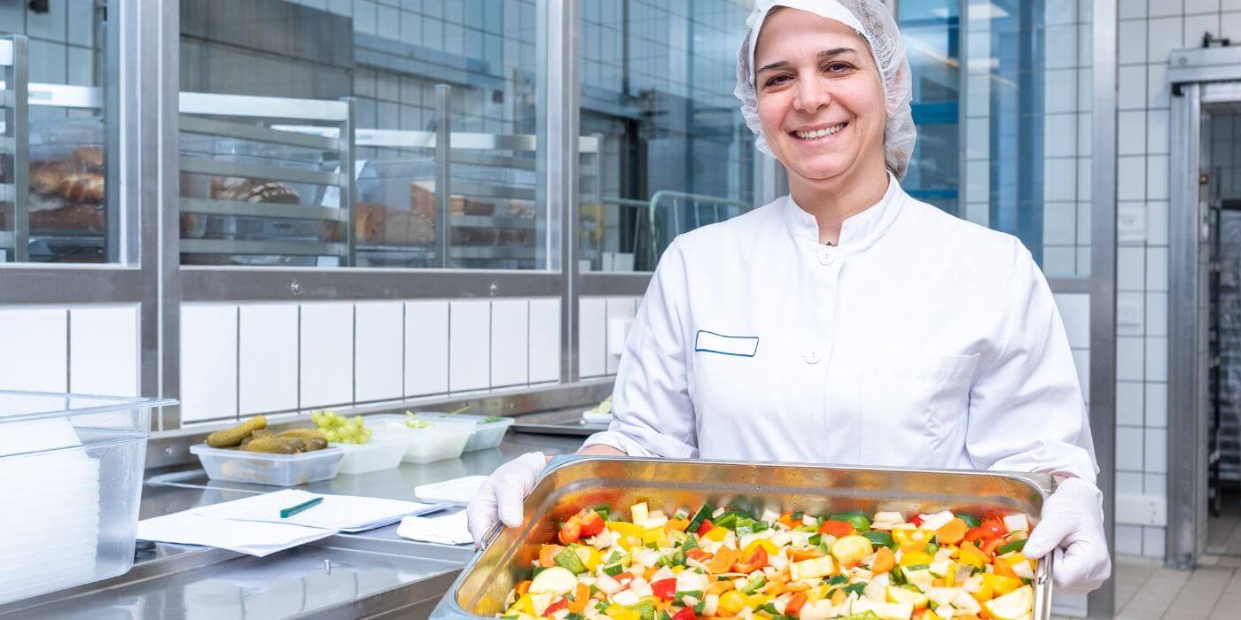 Eine Mitarbeiterin in weißer Arbeitskleidung und Haarnetz hält ein Tablett mit frisch zubereitetem Gemüse in einer Großküche. Steht für Verpflegungsvorteile wie eine Kantine