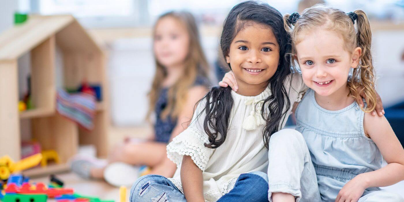 Zwei lachende Kinder sitzen eng beieinander. Ein Mädchen mit dunklen Haaren und ein Mädchen mit blonden Haaren umarmen sich freundlich. Im Hintergrund sind weitere Kinder unscharf zu erkennen