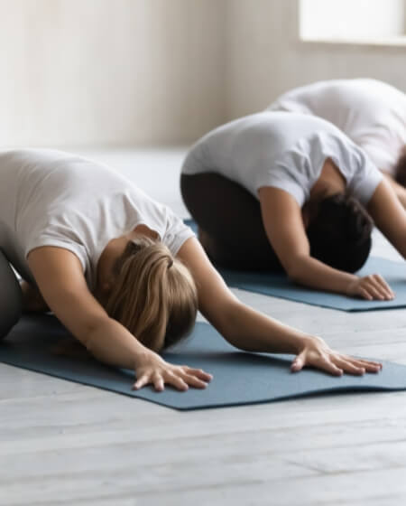 Menschen in sportlicher Kleidung machen eine Yoga-Übung auf Matten in einem hellen Raum. Sie befinden sich in der Position des Kindes (Balasana)