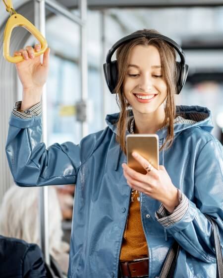 Junge Frau in einer blauen Jacke mit Kopfhörern, die in öffentlichen Verkehrsmitteln steht und lächelnd auf ihr Smartphone schaut. Symbolisiert Mobilitätsvorteile wie ein Jobticket