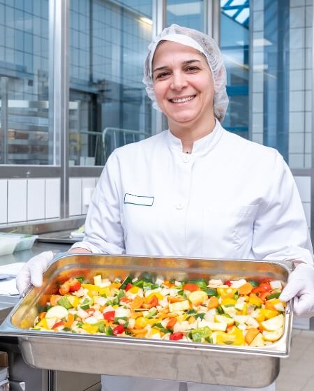 Eine Mitarbeiterin in weißer Arbeitskleidung und Haarnetz hält ein Tablett mit frisch zubereitetem Gemüse in einer Großküche. Steht für Verpflegungsvorteile wie eine Kantine