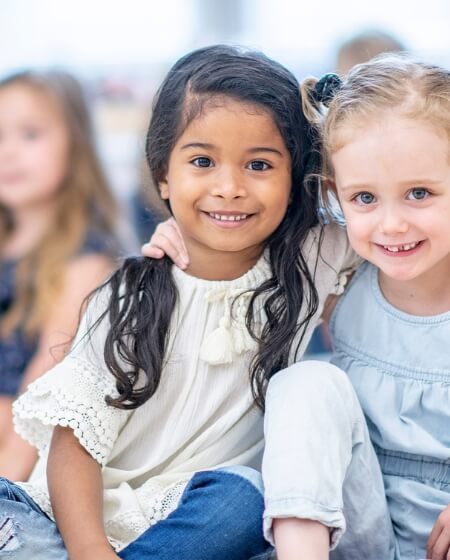 Zwei lachende Kinder sitzen eng beieinander. Ein Mädchen mit dunklen Haaren und ein Mädchen mit blonden Haaren umarmen sich freundlich. Im Hintergrund sind weitere Kinder unscharf zu erkennen