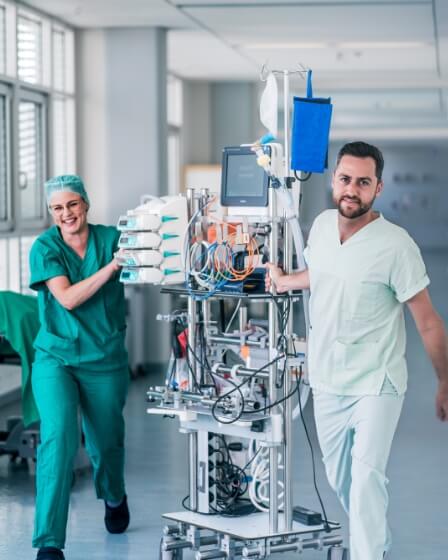 Zwei Personen in grüner medizinischer Kleidung bewegen gemeinsam ein mobiles medizinisches Gerät durch einen Krankenhausflur. Das Gerät ist mit Monitoren, Infusionspumpen und Kabeln ausgestattet. Die Umgebung wirkt technisch und klinisch
