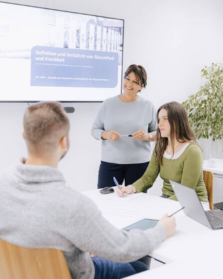 Eine Frau steht lächelnd vor einer Präsentation und erklärt etwas zwei Personen, die an einem Tisch sitzen und Notizen machen. Im Hintergrund ist eine Folie mit dem Titel ‚Definition und Verhältnis von Gesundheit und Krankheit‘ zu sehen