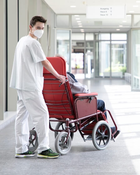 Ein junger Pflegefachhelfer schiebt eine Person in einem Sitzwagen