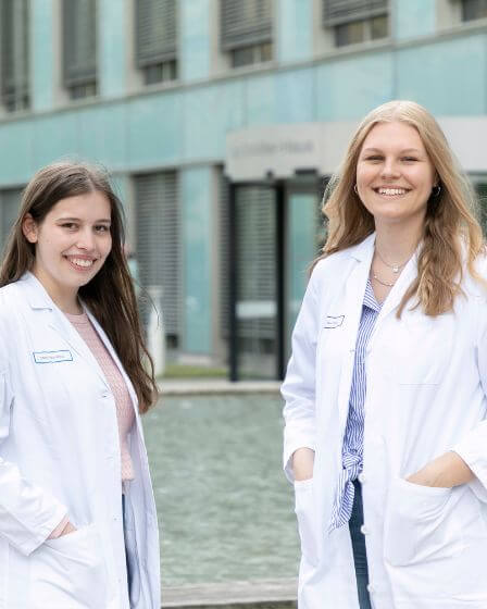 Zwei junge Frauen in weißen Arztkitteln stehen vor einem modernen Gebäude mit großen Glasfenstern.