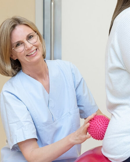 Eine Hebamme in hellblauer Arbeitskleidung unterstützt eine schwangere Frau, die auf einem roten Gymnastikball sitzt. Die Hebamme hält einen Massageball in der Hand und führt eine entspannende Übung durch