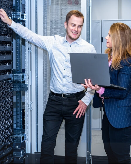Ein Mann und eine Frau stehen in einem Serverraum. Der Mann zeigt auf ein Serverrack, während die Frau mit einem Laptop in der Hand etwas erklärt