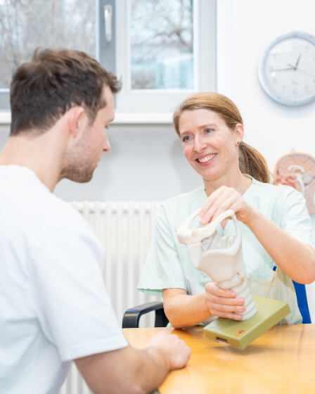 Eine Logopädin in medizinischer Kleidung zeigt einem Mann ein anatomisches Modell des Kehlkopfes und erklärt dessen Funktion in einem hellen Raum mit medizinischen Anschauungsmaterialien