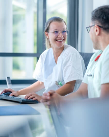 Eine Medizinische Fachangestellte in weißer Arbeitskleidung sitzt an einem Schreibtisch und lächelt ihren Kollegen an, der ebenfalls in medizinischer Kleidung neben ihr sitzt