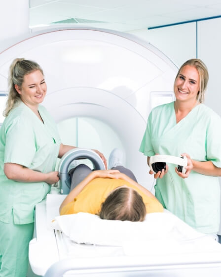 Zwei MTRAs in grüner medizinischer Kleidung bereiten einen Patienten für eine Untersuchung in einem MRT-Gerät vor und lächeln freundlich in die Kamera