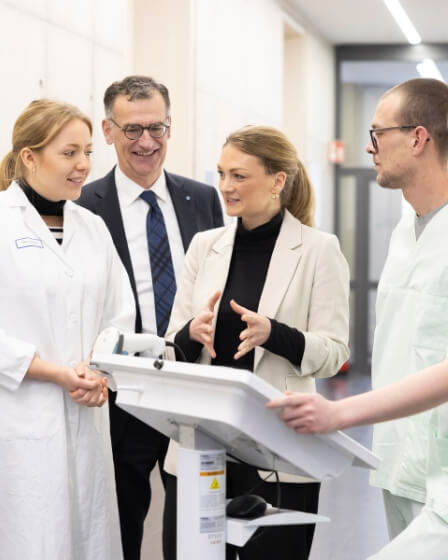 Eine Gruppe von vier Personen, darunter zwei Frauen in weißen Laborkitteln, ein Mann im Anzug und eine Frau in einem beigen Blazer, stehen vor einem medizinischen Gerät und unterhalten sich angeregt