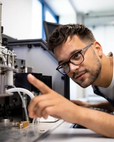Ein Mechatroniker mit Brille und Arbeitskleidung inspiziert konzentriert eine technische Maschine und zeigt mit dem Finger auf ein Bauteil