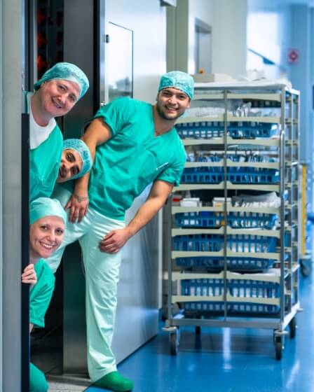 Ein fröhliches Team von medizinischem OP-Personal in grüner OP-Kleidung schaut lachend aus einer Tür. Im Hintergrund sind medizinische Gerätewagen zu sehen