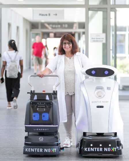 Eine Reinigungskraft steht mit zwei modernen Reinigungsrobotern in einem hellen Innenbereich eines Gebäudes