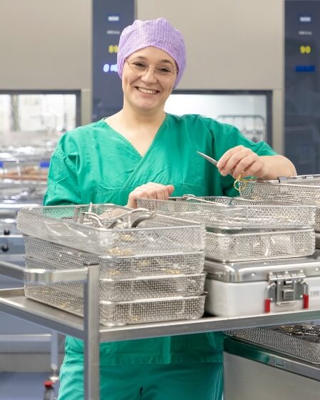 Eine Frau in grüner Arbeitskleidung und lilafarbener Haube steht in der ZSVA. Sie arbeitet an medizinischen Instrumenten, die in Metallbehältern auf einem Rollwagen liegen, und lächelt in die Kamera