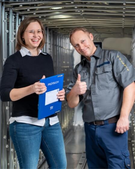 Eine Ingenieurin und ein Techniker stehen in einem technischen Raum mit großen Metallanlagen. Die Frau hält eine blaue Mappe mit der Aufschrift „Prüfbuch“ und lächelt, während der Mann mit Daumen hoch in die Kamera blickt