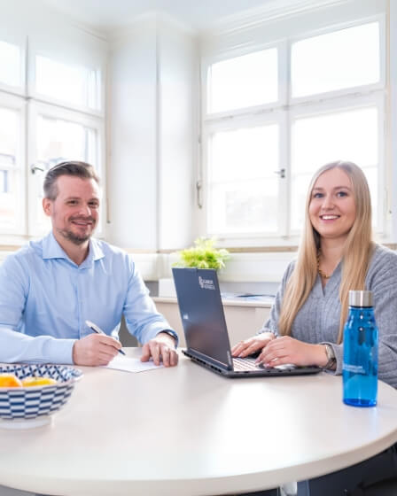 Ein Mann und eine Frau sitzen an einem Tisch in einem Büro mit großen Fenstern. Beide lächeln, während die Frau an einem Laptop arbeitet und der Mann Notizen macht