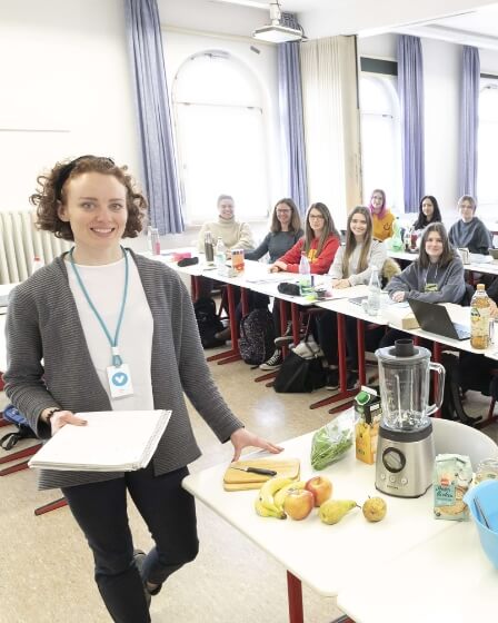 Eine Lehrkraft steht lächelnd vor einer Gruppe von Auszubildenden in einem Schulungsraum. Auf einem Tisch vor ihr liegen Obst, ein Mixer und weitere Zutaten. Sie hält Unterlagen in der Hand und trägt ein Namensschild. Im Hintergrund sitzen mehrere Personen, die aufmerksam zuhören