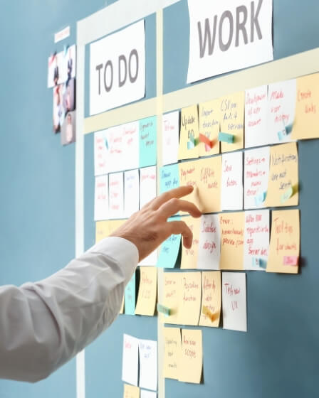 Eine Hand zeigt auf ein Kanban-Board mit farbigen Haftnotizen, das in die Spalten 'To Do' und 'Work' unterteilt ist. Das Bild symbolisiert Organisation, Teamarbeit und Aufgabenmanagement