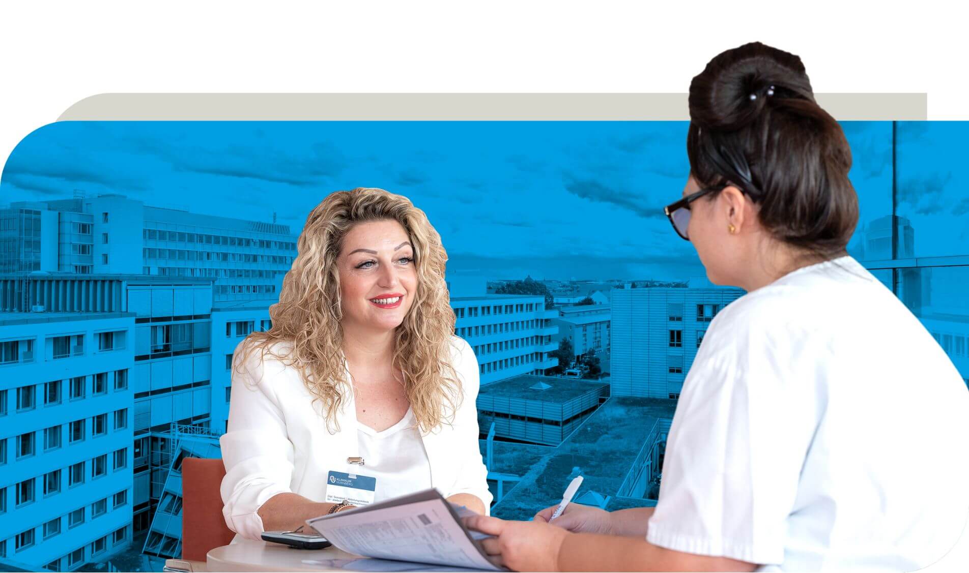 Zwei Frauen sitzen an einem Tisch in einem Gespräch. Eine Frau trägt ein weißes Oberteil und hält Dokumente, während die andere Frau in Arbeitskleidung Notizen macht. Das Bild stellt ein Beratungsgespräch dar