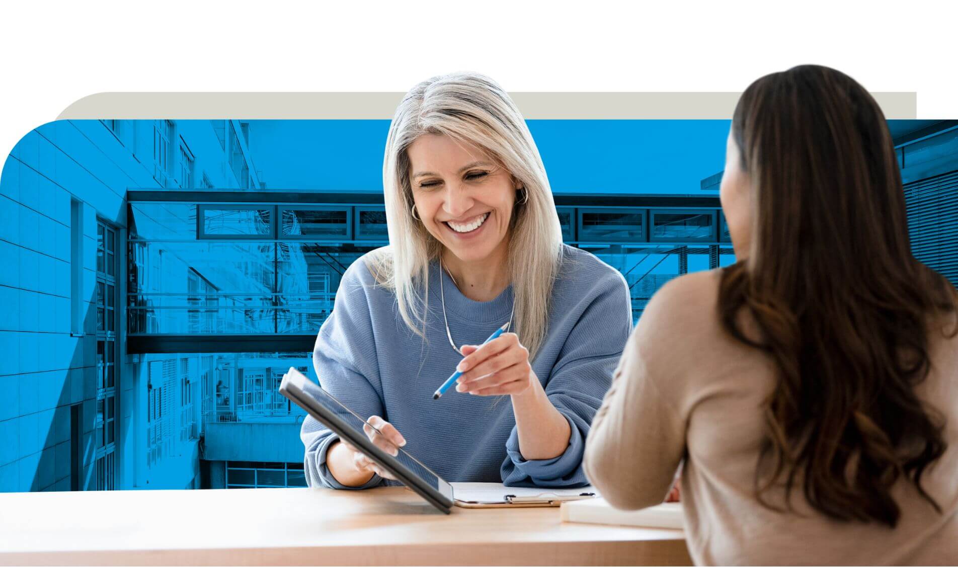 Eine Frau in einem blauen Pullover bespricht mit einer anderen Person Details an einem Tisch. Sie hält einen Stift und ein Tablet, während sie etwas erklärt