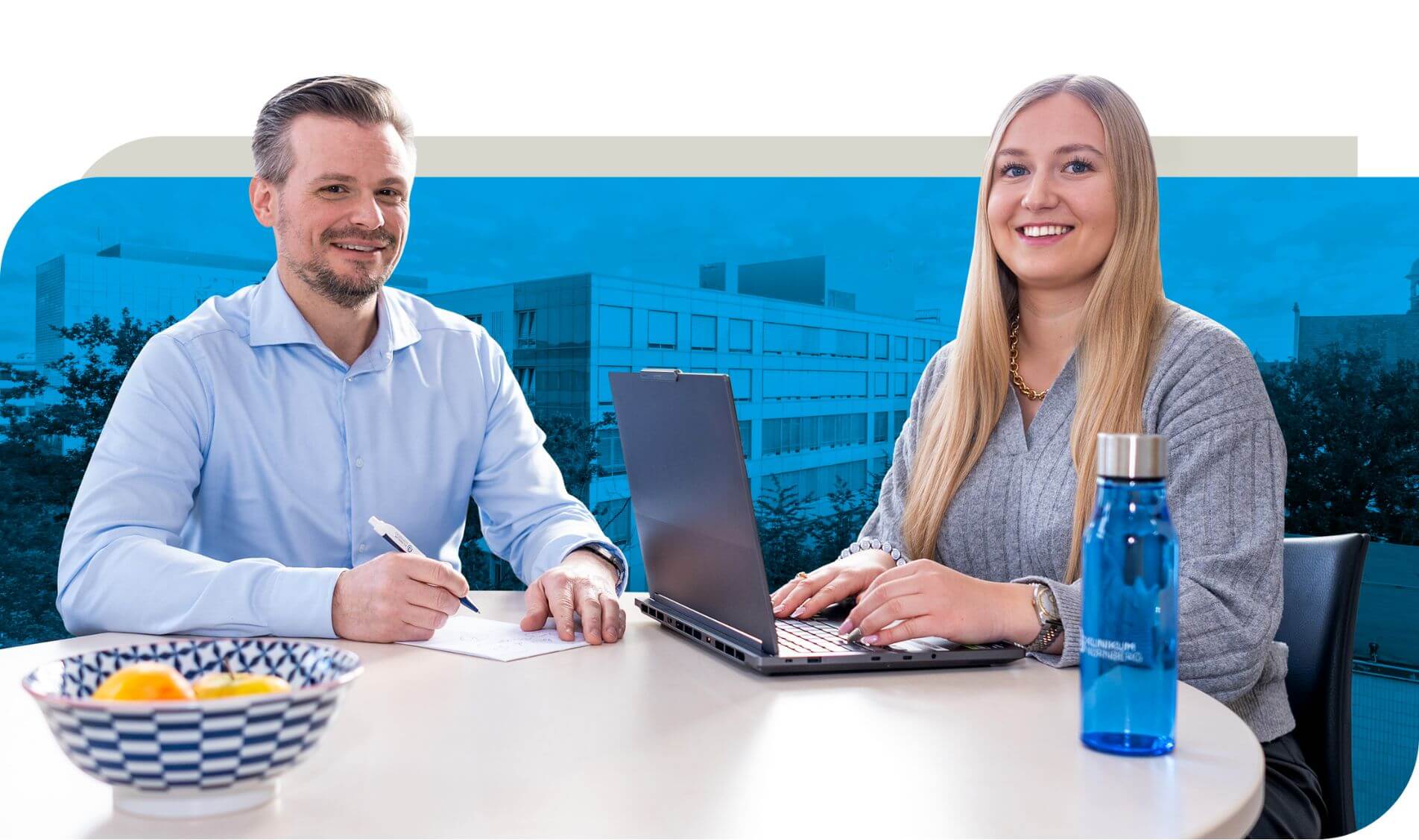 Zwei Personen sitzen an einem Tisch. Eine Person schreibt Notizen auf Papier, während die andere an einem Laptop arbeitet. Auf dem Tisch stehen eine blaue Wasserflasche und eine Schale mit Obst. Im Hintergrund ist ein Bürogebäude zu erkennen